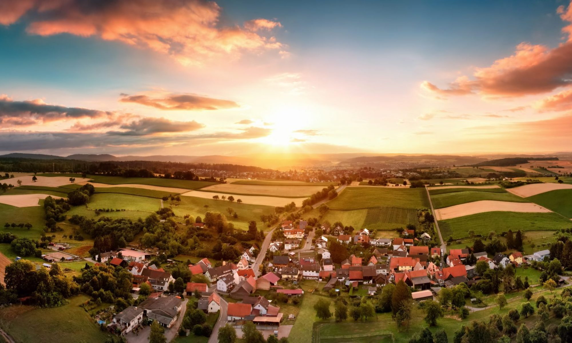 Aerial Panorama eines Dorfes umgeben von Feldern bei Sonnenaufgang, mit schönen bunten Himmel und warmes Licht